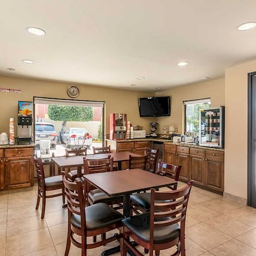 The image shows a small dining area with tables and chairs, a coffee and beverage station, a TV, and a window overlooking the outside.