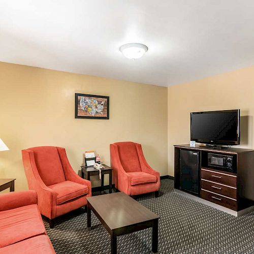 A hotel living area with red chairs, a sofa, a TV on a cabinet, a coffee table, a lamp, and an adjacent bedroom visible through a doorway.