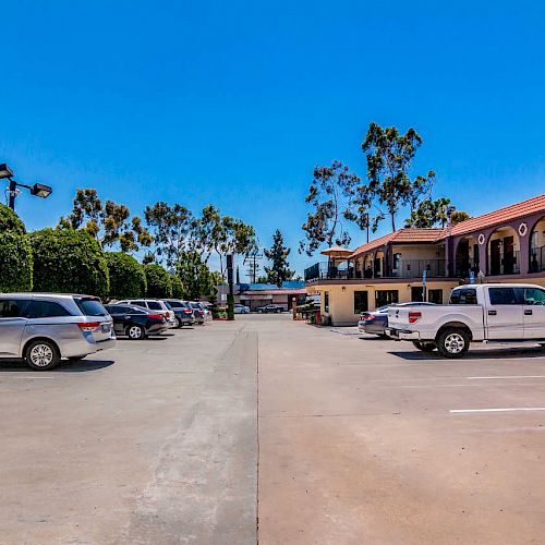 This image shows an outdoor parking lot with several vehicles parked beside a two-story building with balconies, under a clear blue sky.