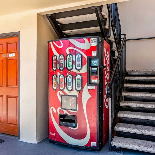 A red vending machine is placed next to a staircase outside a building, with an orange door marked 