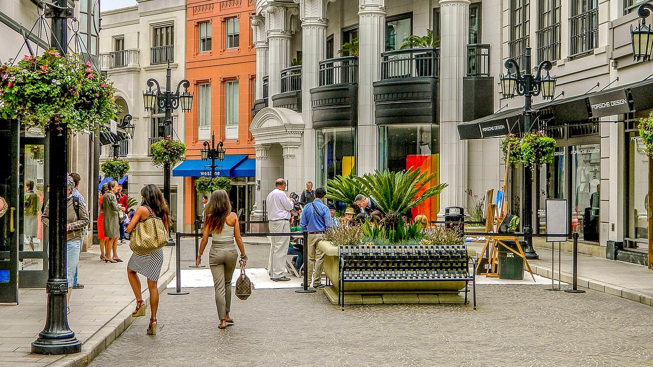 A bustling outdoor shopping area with people walking, ornate buildings, potted plants, and decorative street lamps under a partly cloudy sky.