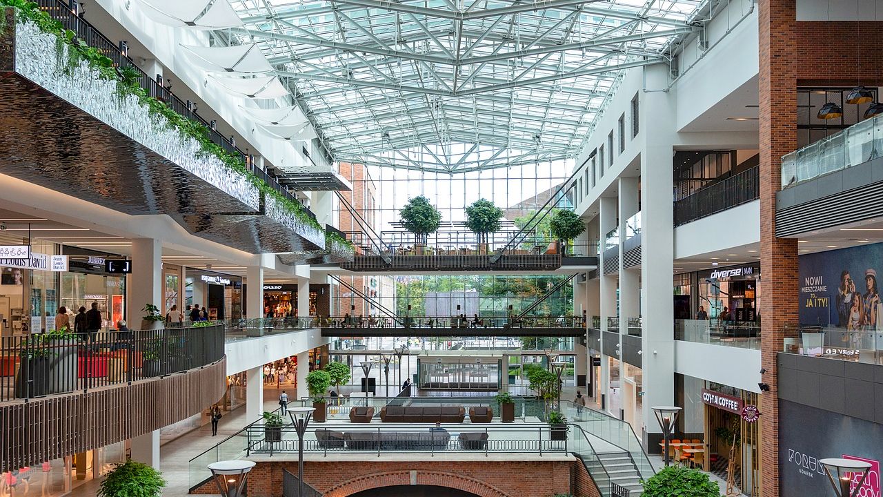 A modern, spacious shopping mall with multiple levels, a large glass ceiling, various shops, potted plants, and numerous seating areas.