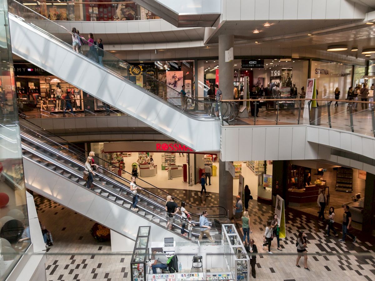 A bustling shopping mall with escalators, various shops, and people walking and browsing around.