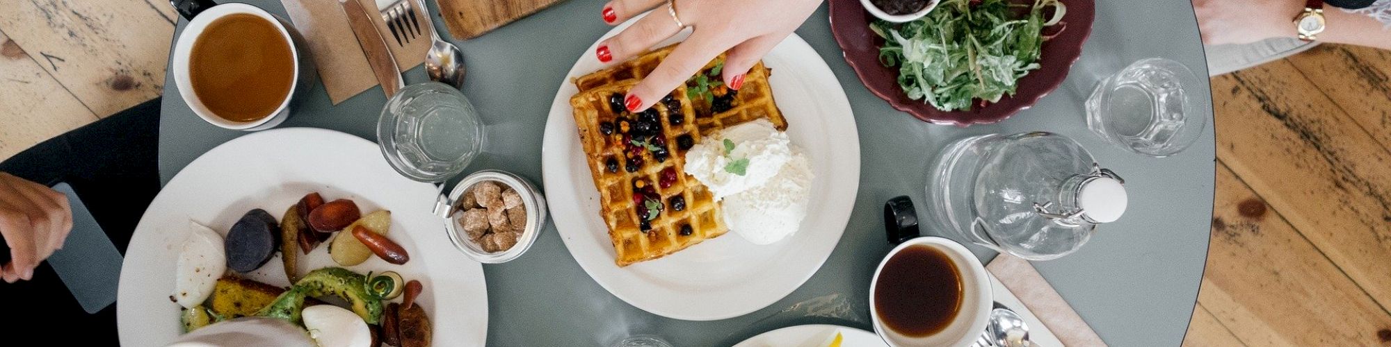 This is an overhead view of a dining table with various dishes, including waffles, salad, coffee, and utensils, with people reaching for food.