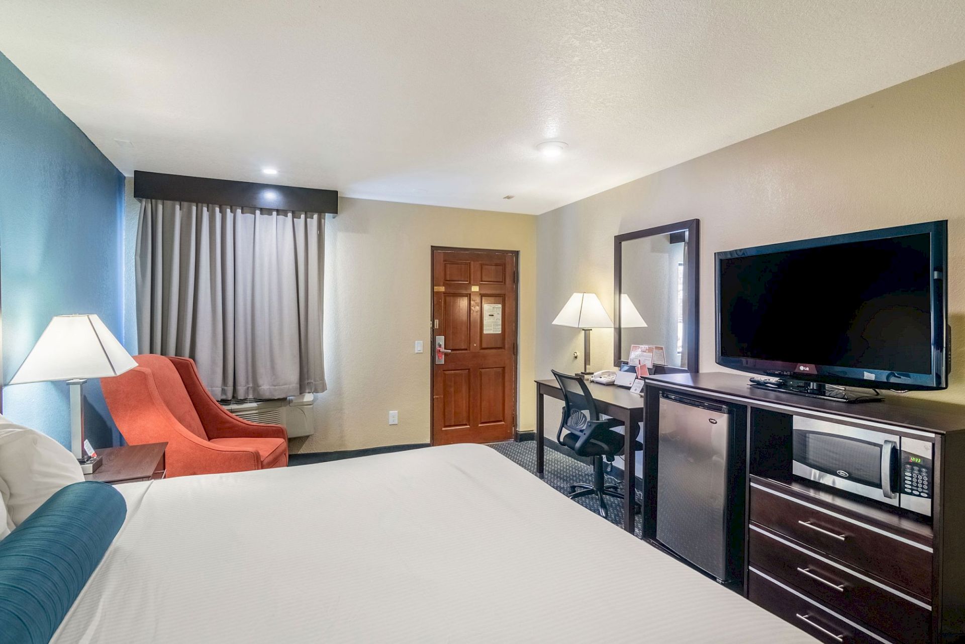 This image shows a hotel room with a bed, an orange chair, lamps, a desk with a chair, a large TV, a mini-fridge, and a microwave.