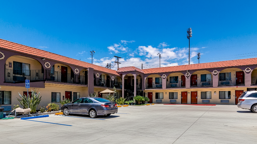 The image shows a two-story motel with parked cars in front, featuring a courtyard and a clear blue sky above.