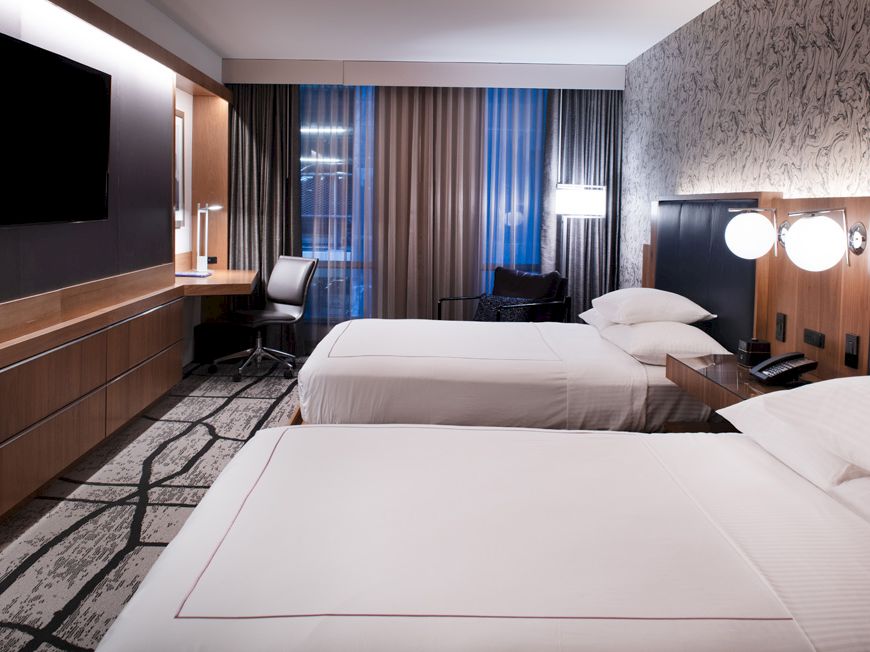 A modern hotel room with two beds, a wall-mounted TV, a desk with a chair, lamps on the nightstands, and floor-to-ceiling windows with curtains.