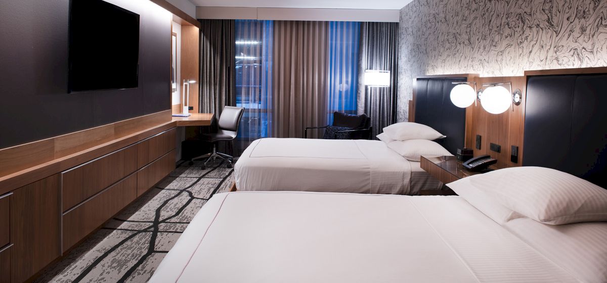 A modern hotel room with two beds, a wall-mounted TV, a desk with a chair, lamps on the nightstands, and floor-to-ceiling windows with curtains.