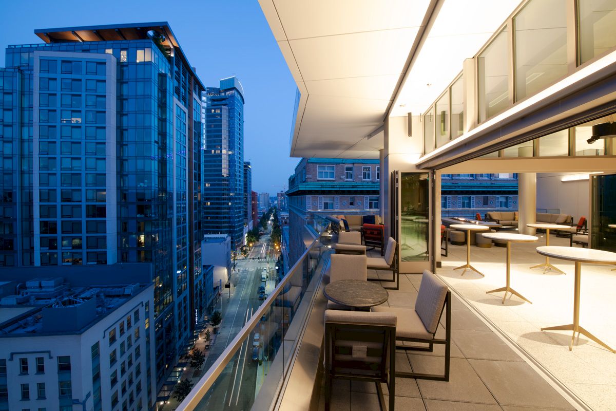 The image shows a modern balcony with seating and tables, overlooking a cityscape with tall buildings and a lit street below at dusk.
