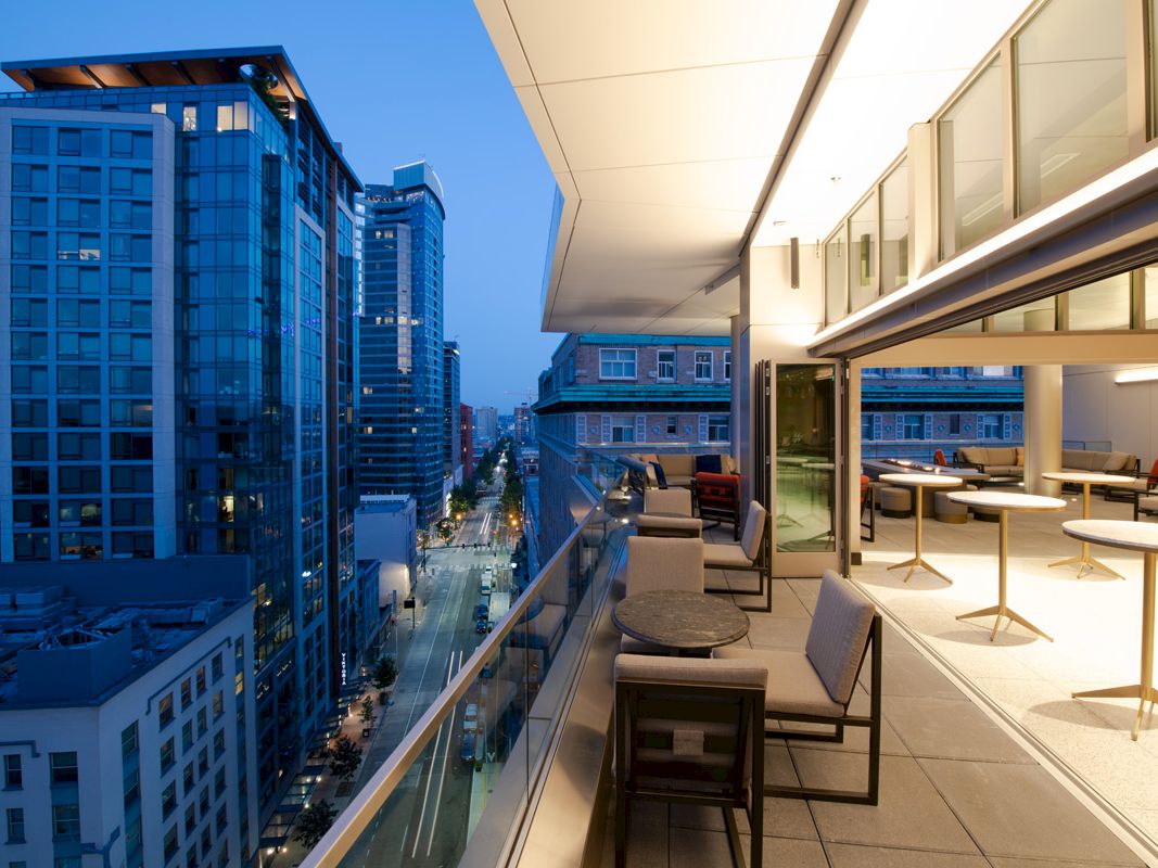 The image shows a modern balcony with seating and tables, overlooking a cityscape with tall buildings and a lit street below at dusk.