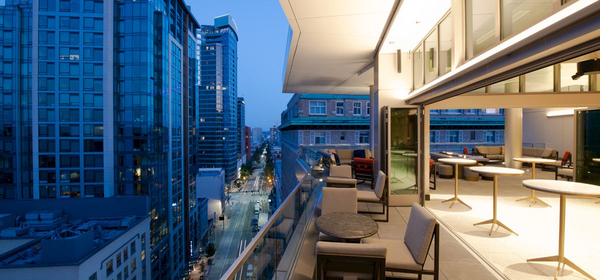 The image shows a modern balcony with seating and tables, overlooking a cityscape with tall buildings and a lit street below at dusk.