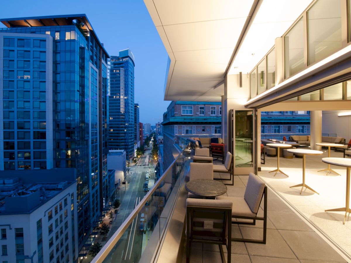 A rooftop balcony with seating areas overlooks a city street, flanked by modern high-rise buildings during dusk, with city lights illuminating.