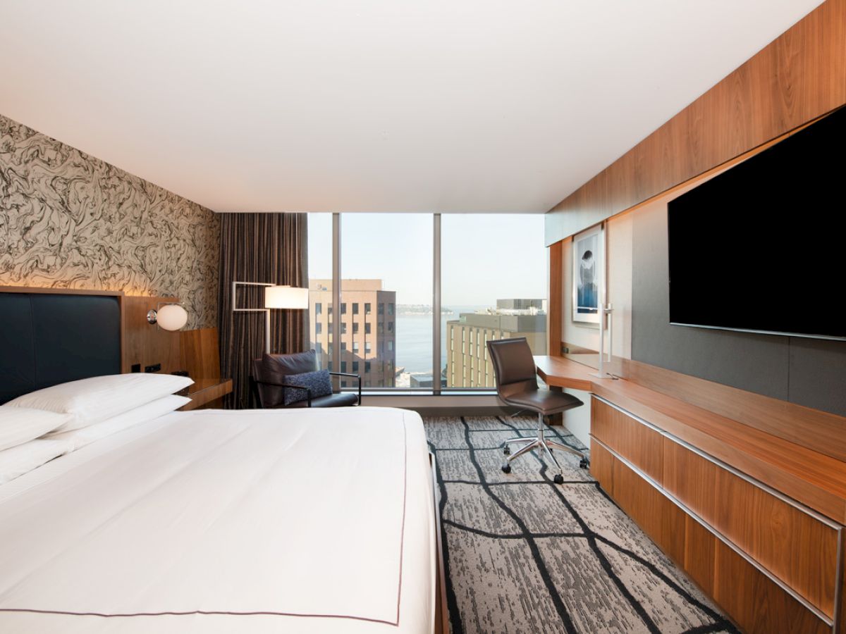 A modern hotel room with a large bed, desk, chair, TV, and a window offering a city view. The room features wooden accents and a stylish design.