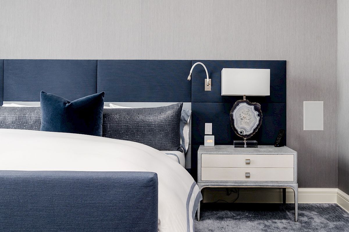 A modern bedroom with a blue-themed bed, gray walls, a nightstand with a lamp, and a decorative object on top, ending the sentence.