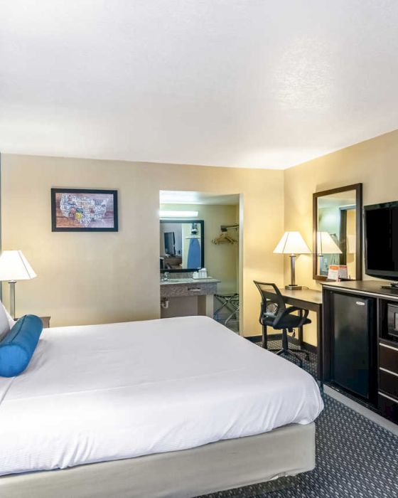 The image shows a hotel room with a large bed, two bedside lamps, a TV on a dresser, a desk, a chair, and a bathroom in the background.